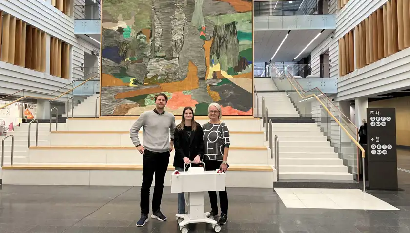 Tre personer står i en modern byggnad framför en stor, färgstark konstvägg. De står på en bred trappa med träbänkar och glasräcken på sidorna. Personen i mitten håller i en vit medicinsk apparat på hjul. De är klädda i en blandning av formella och avslappnade kläder. Bakgrunden visar en öppen interiör med skyltar och butiker.