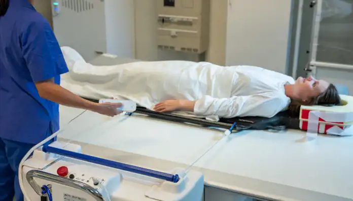 En patient i en vit sjukhusrock ligger på en medicinsk behandlingsbädd, stödd av en specialanpassad huvudkudde. En sjukvårdspersonal i blå uniform håller en fjärrkontroll och styr en medicinsk apparat. Rummet har en klinisk miljö med medicinsk utrustning och paneler i bakgrunden.