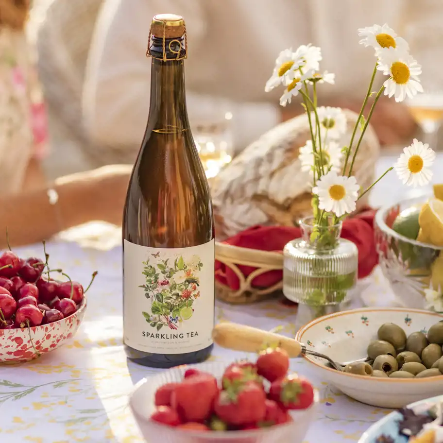 En flaska med etiketten "Sparkling Tea" står i centrum på ett somrigt dukat bord. Runt flaskan finns färska jordgubbar, körsbär, gröna oliver, ett bröd i bakgrunden och en liten vas med prästkragar. Bilden förmedlar en känsla av en solig picknick eller en trädgårdsfest.