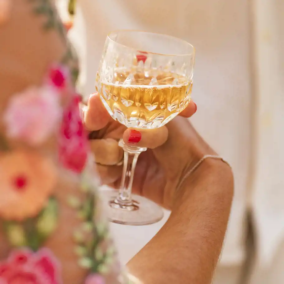 En närbild på en hand med rödmålade naglar som håller ett elegant kristallglas fyllt med en gyllene mousserande dryck. I bakgrunden skymtar en ljus klädsel och färgglada broderade blommor, vilket ger en somrig och festlig känsla.