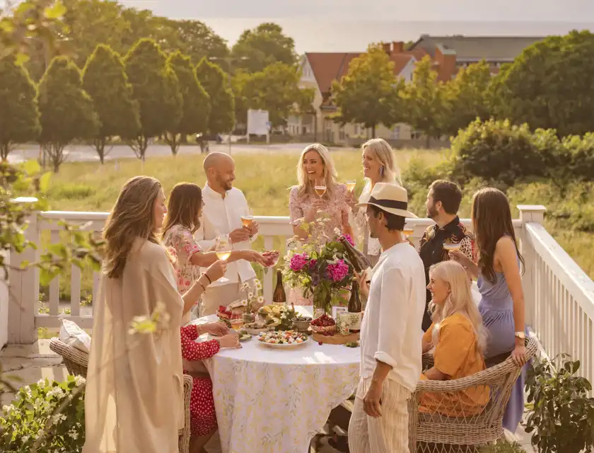 Ett glatt sällskap på elva personer samlade runt ett somrigt dukat bord på en solig altan. De håller glas med dryck och skrattar tillsammans. Bordet är dekorerat med färska blommor, frukt och lätta sommarrätter. I bakgrunden syns grönskande träd, hus och ett glimtande vatten.