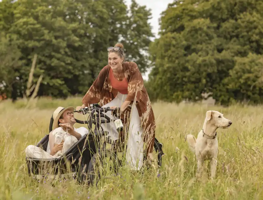En kvinna skrattar och kör en lådcykel genom en sommaräng, medan en man sitter i lådan med ett glas i handen och ler upp mot henne. En ljus hund går bredvid i det höga gräset. I bakgrunden syns gröna träd och en molnig himmel.