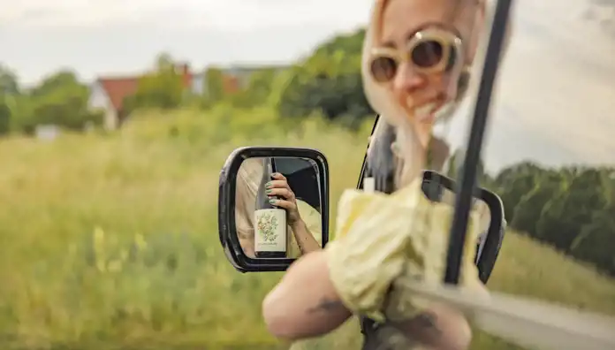 En kvinna med solglasögon ler medan hon lutar sig ut genom fönstret på en bil. I sidospegeln syns hennes hand hålla upp en flaska Sparkling Tea med en blomillustration på etiketten. I bakgrunden syns en somrig äng med grönt gräs och suddiga hus.