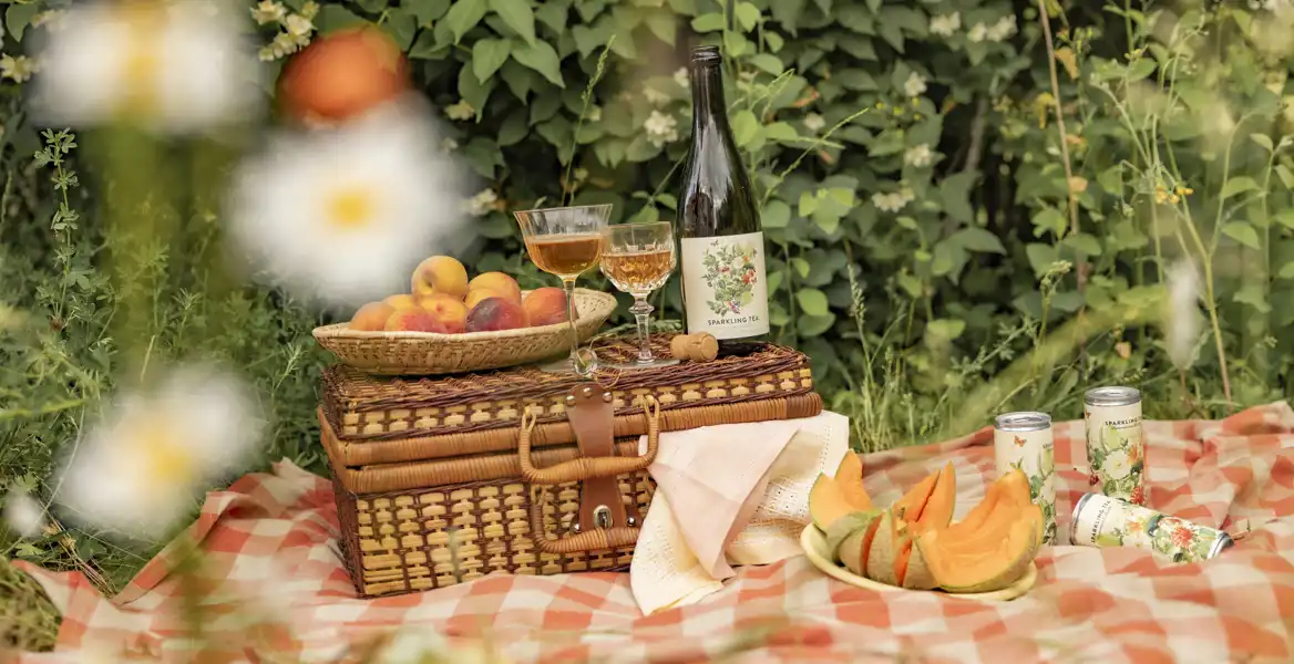 En picknickscen med en rutig filt på gräset. På filten står en flätad picknickkorg med en skål persikor ovanpå, två glas med gyllene dryck, en flaska Sparkling Tea och några servetter. Till höger syns en tallrik med klyftad melon och tre burkar Sparkling Tea. I bakgrunden frodig grönska och blommor.