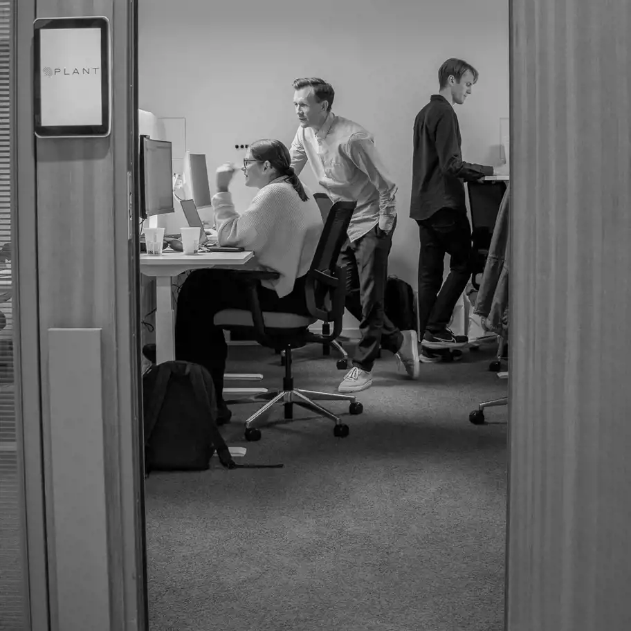 Tre personer arbetar fokuserat vid datorer i ett modernt kontorsrum hos företaget Plant. Bilden förmedlar ett kreativt och produktivt arbetsklimat.