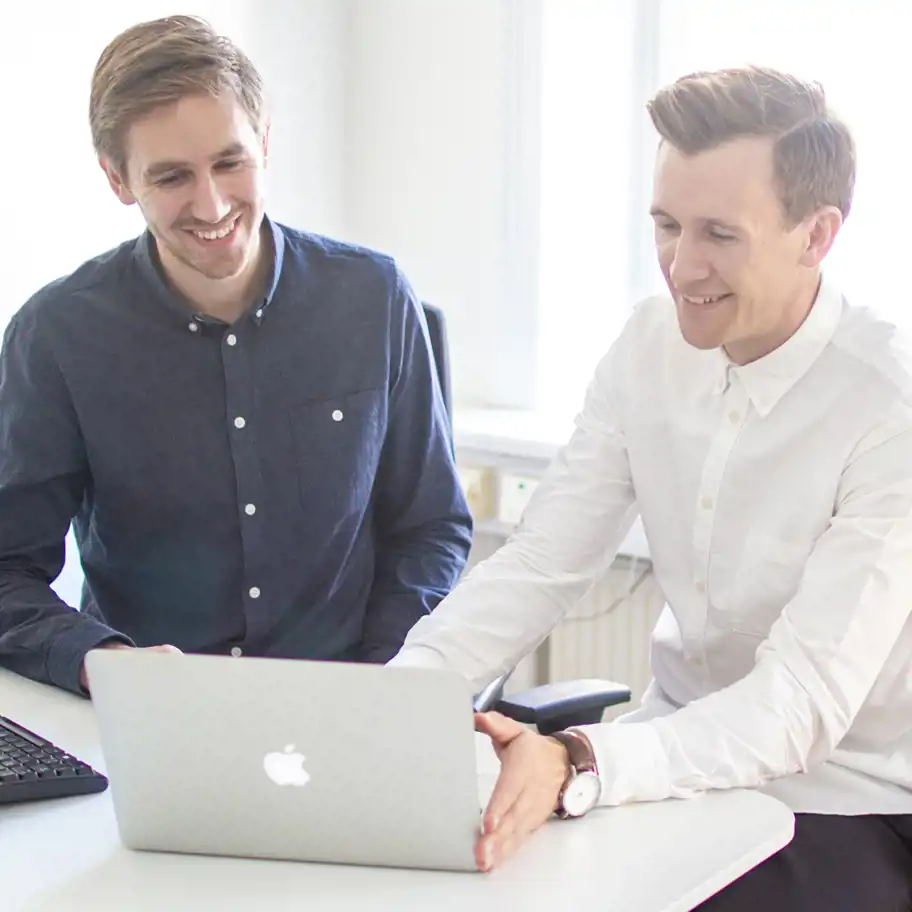 Två unga män sitter vid ett skrivbord och arbetar tillsammans framför en laptop i ett ljust kontorsrum. De ler och ser engagerade ut.