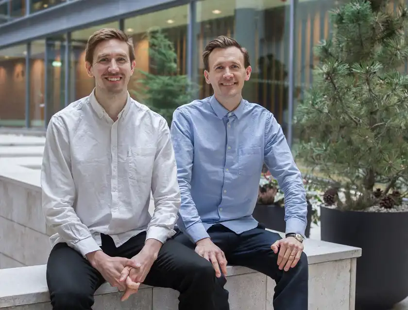 Två leende män som är företagare sitter utomhus i en modern kontorsmiljö, omgivna av grönska.