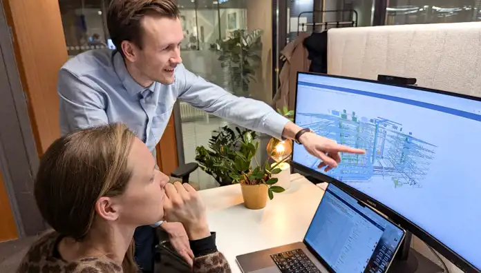 En man pekar på en teknisk ritning på en datorskärm medan en kvinna lyssnar och tittar koncentrerat. Bilden visar ett innovativt arbetsmoment i en modern kontorsmiljö.