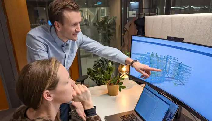 En man pekar på en teknisk ritning på en datorskärm medan en kvinna lyssnar och tittar koncentrerat. Bilden visar ett innovativt arbetsmoment i en modern kontorsmiljö.