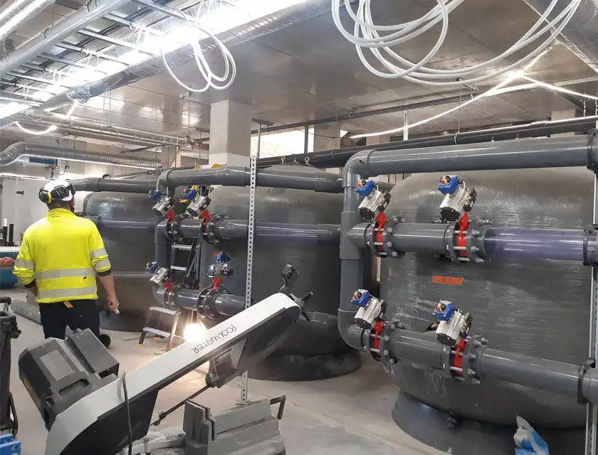 Industrimiljö med stora filtertankar och rörsystem. En person i gul varseljacka och hörselskydd står och inspekterar installationen. Flera ventiler och kopplingar är synliga på rören, och taket är fyllt med hängande kablar och ventilationsrör. I förgrunden syns en maskin märkt "poolwater".
