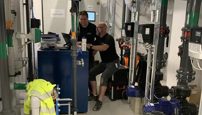 Två män i svarta tröjor med "poolwater"-logotyp arbetar vid en dator i en teknisk anläggning. De sitter och står vid ett blått skåp omgivet av rör, ventiler och pumpar. En gul varseljacka hänger på ett rör i förgrunden. Bakom dem syns en skärm med företagets logotyp och mappar staplade på skåpet.