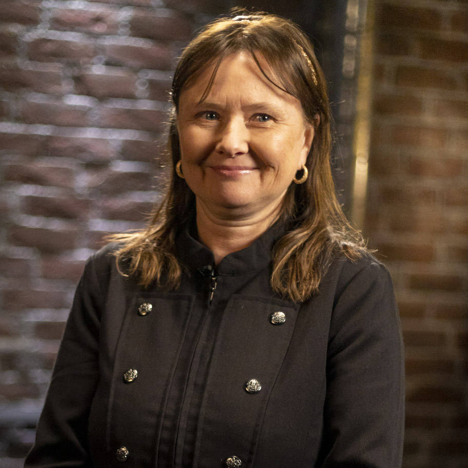 En kvinna med axellångt brunt hår och guldörhängen står i en mörk miljö med tegelvägg i bakgrunden. Hon bär en svart jacka med dekorativa knappar och ser nöjd ut. I övre vänstra hörnet syns texten "Draknästet" och i övre högra hörnet SVT:s logotyp.