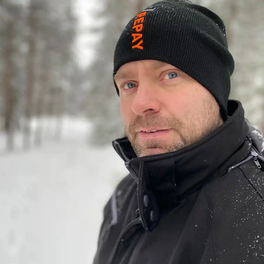 En man med kort skägg och blå ögon står ute i ett vintrigt landskap med snötäckta träd i bakgrunden. Han bär en svart mössa med orange text "REPAY" och en svart vinterjacka med hög krage och snöflingor på axlarna.