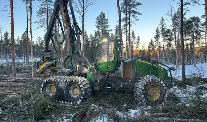 En stor grön skördare från John Deere står i en avverkad skog med snötäckta grenar och trädstammar på marken. Maskinen har en lång kran med ett skördaraggregat i gul och svart färg. Hjulen är försedda med kedjor för bättre grepp i den snöiga terrängen. Solen går ner i bakgrunden och skapar ett varmt ljus mellan de kvarvarande träden.
