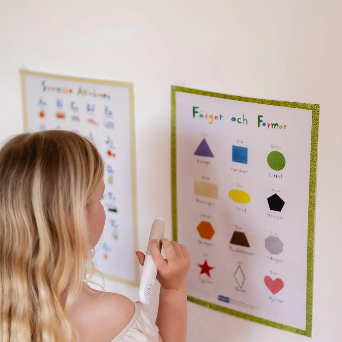 Ett barn använder Readioos läspenna för att peka på en plansch med färger och former på väggen. Pennan spelar upp ljud kopplade till bilderna.