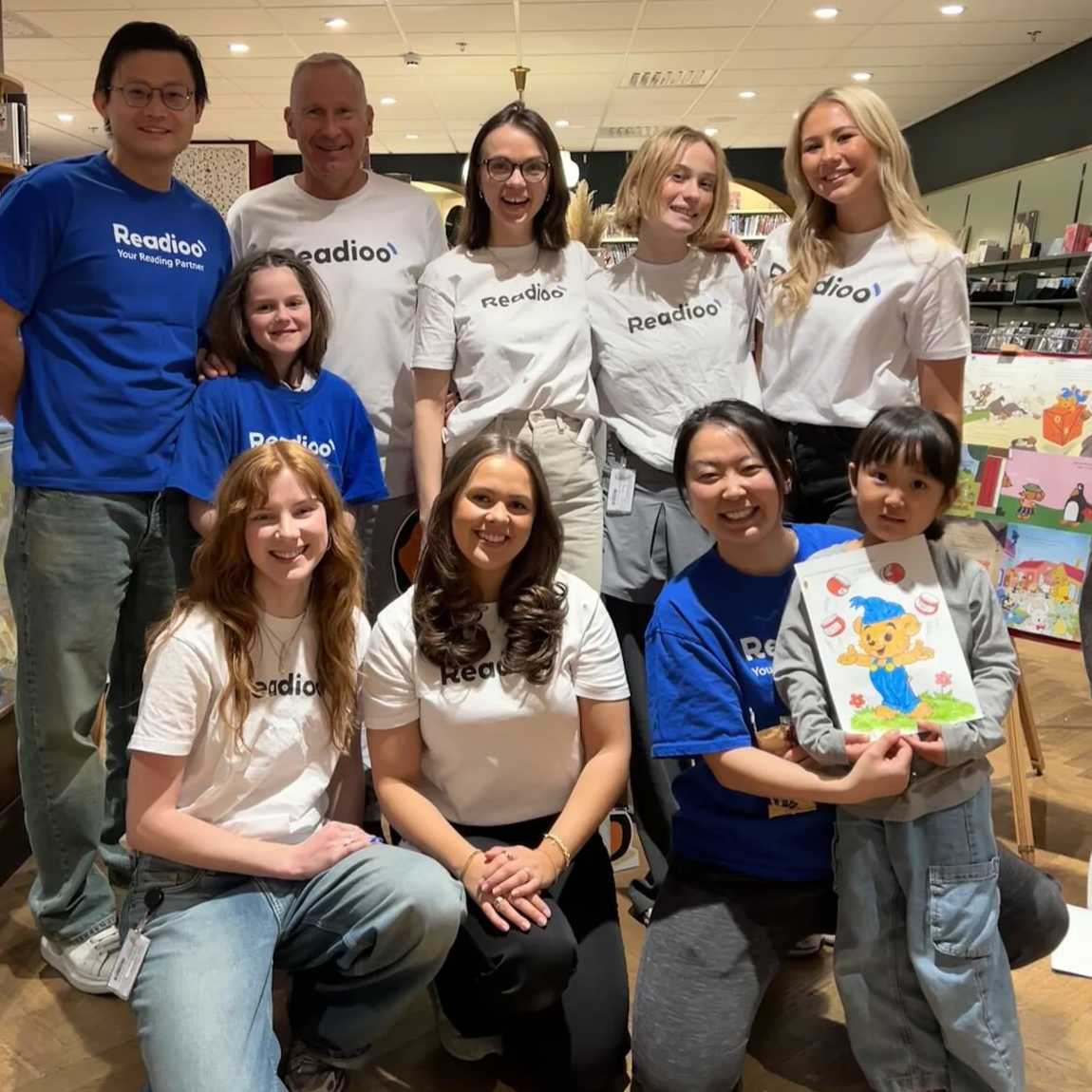 Gruppbild på Readioos team som ler mot kameran. Några bär vita och blå tröjor med Readioo-logotypen. En kvinna håller i ett barn som visar en teckning med Bamse.