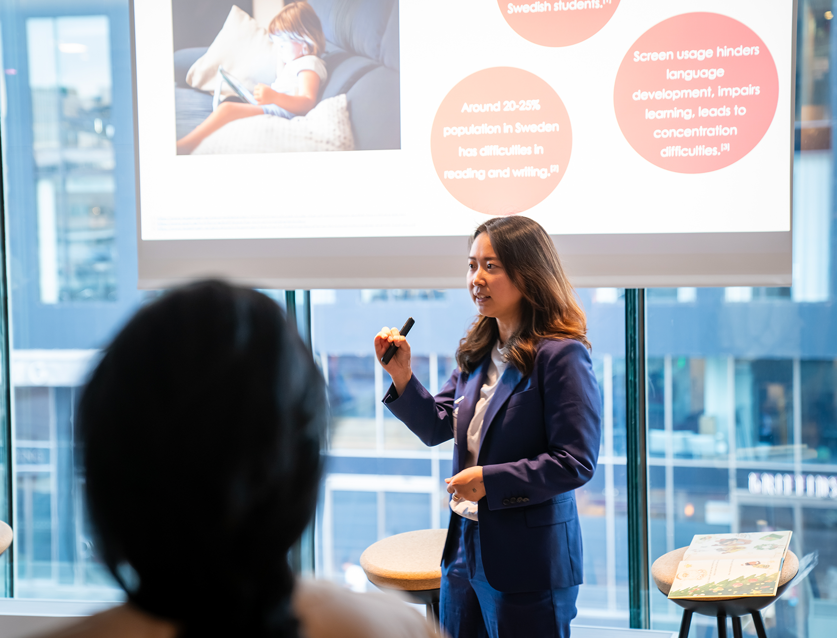 Inga Li, medgrundare till Readioo, håller en presentation framför en projektorduk med bilder och text. Hon talar inför en publik och visar en bild med en pekare.