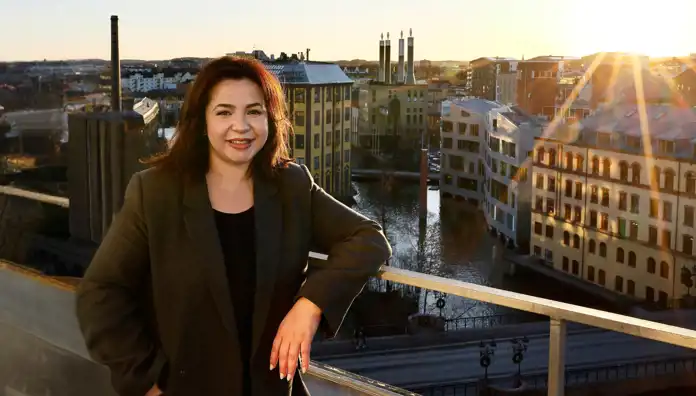 En kvinna med axellångt mörkt hår och ett vänligt leende står lutad mot ett räcke med en stadsmiljö i bakgrunden. Hon bär en mörkgrå kavaj över en svart topp. Solen lyser lågt och skapar en varm ljuseffekt över byggnaderna och vattnet, där både moderna och äldre byggnader speglas.
