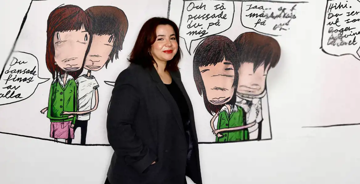 En kvinna med axellångt mörkt hår och en mörkgrå kavaj står framför en stor, tecknad serieillustration på en vit vägg. Hon ler och har en avslappnad hållning. Serieillustrationen föreställer två personer i en omfamning, med pratbubblor fyllda med handskriven text på svenska.