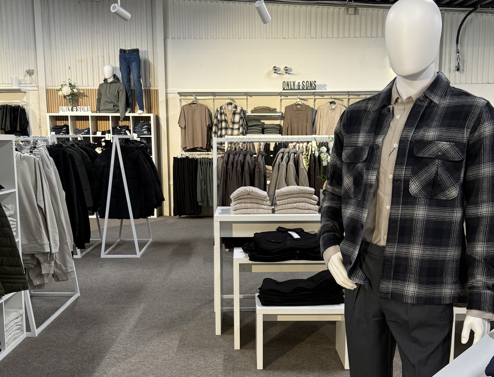 Klädavdelning i Ronneby Outlet med herrkläder upphängda på ställ och vikta på bord. En skyltdocka i rutig skjortjacka står i förgrunden.