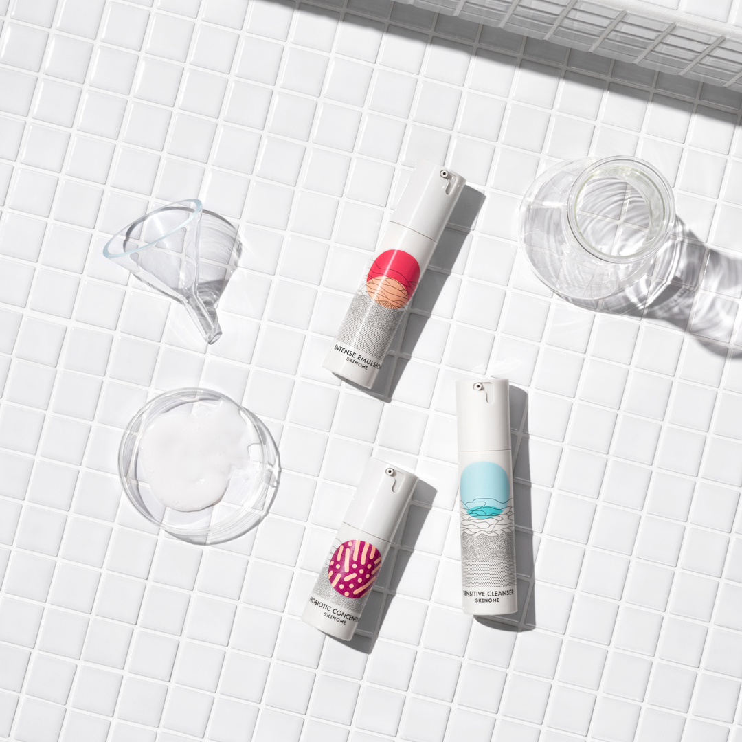 Tre hudvårdsprodukter från Skinome placerade på vit kakelbakgrund, tillsammans med en glaskolv, tratt och en petri-skål med vit kräm.