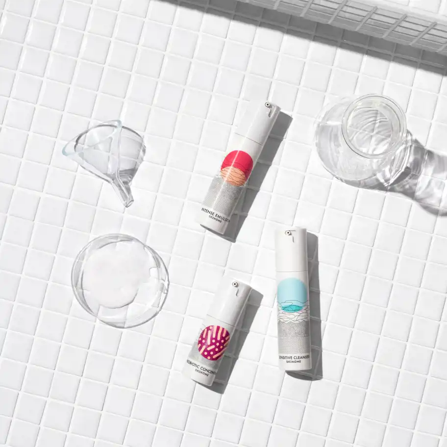 Tre hudvårdsprodukter från Skinome placerade på vit kakelbakgrund, tillsammans med en glaskolv, tratt och en petri-skål med vit kräm.