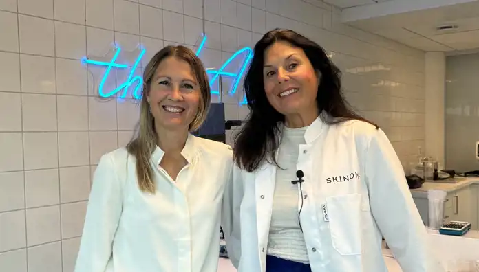 Två kvinnor står i ett ljust labbmiljö. Den ena bär en vit blus och den andra en vit rock med texten "Skinome". Båda ler mot kameran.