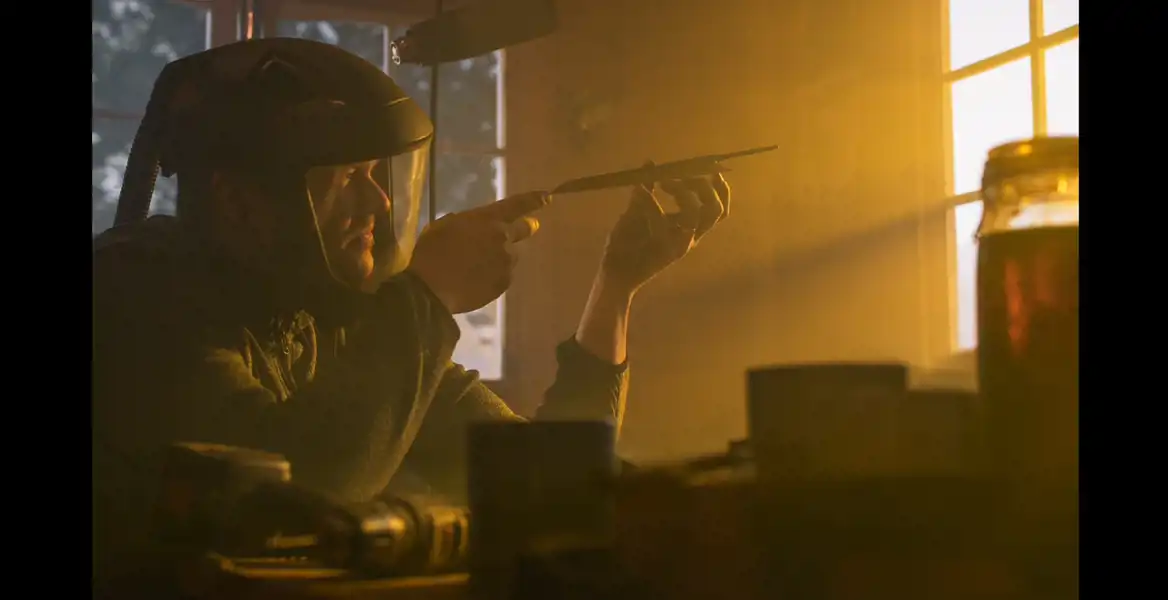 En man, Mattias Lundberg grundare av Steel by Lundbergs, i sin smedja iförd skyddsutrustning och granskar noggrant en kniv i motljus vid ett arbetsbord, med verktyg utspridda omkring sig.