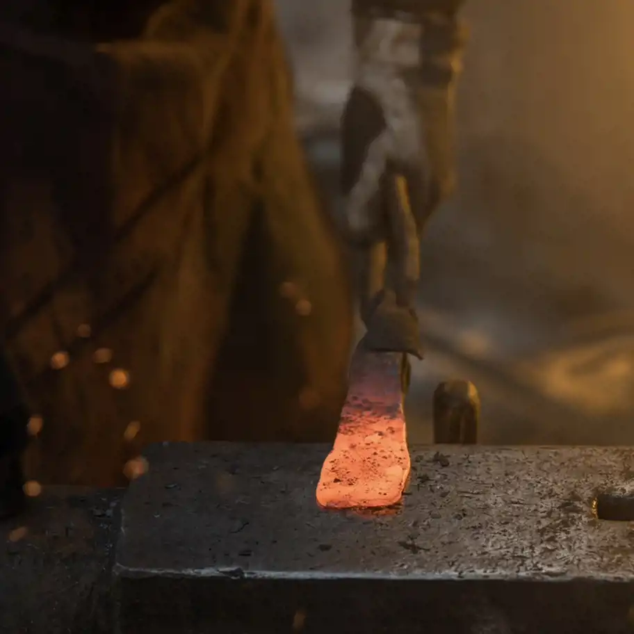 Ett glödande metallstycke hålls fast med tång ovanpå ett städ, redo att smidas vidare. Bilden fångar kraften i hantverk och skapande.