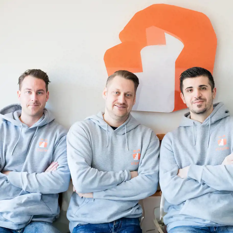 Tre män i grå huvtröjor med Stepler-logotypen poserar leende framför en vägg med en del av företagets logga i bakgrunden – en bild som visar teamet bakom hälsoappen Stepler, som uppmuntrar rörelse genom belöningar och skapar hållbara vanor med hjälp av digital innovation.