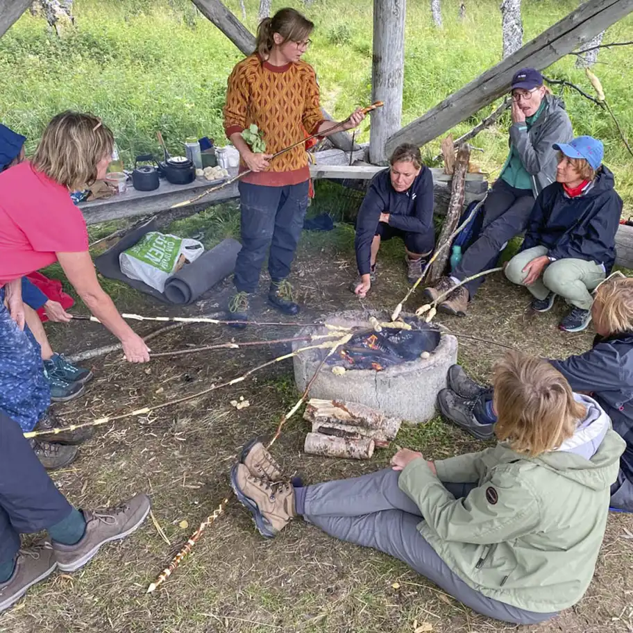 En grupp människor sitter och står runt en öppen eld under ett vindskydd i skogen. De grillar pinnbröd över elden med långa pinnar och verkar vara engagerade i samtal. Runt omkring syns ryggsäckar, liggunderlag och kokkärl, vilket antyder att de är på friluftsutflykt eller kurs i naturen.