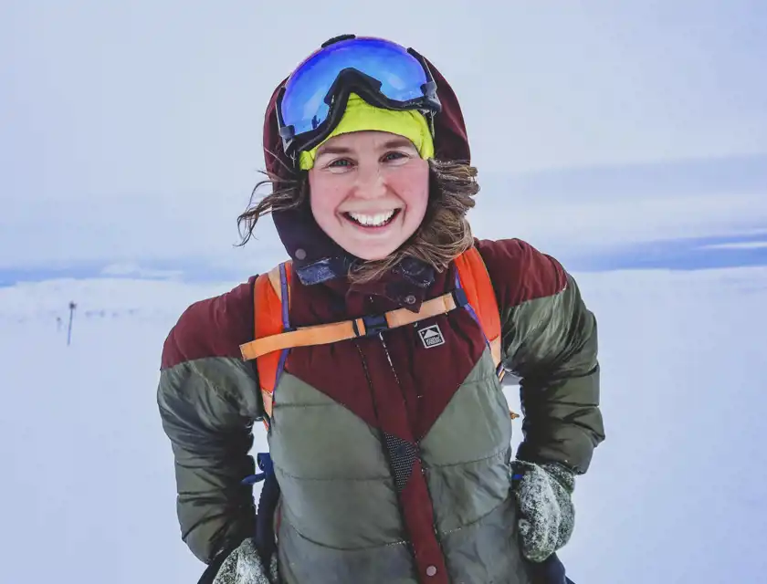 En leende kvinna iklädd vinterkläder står utomhus i ett snötäckt landskap. Hon har på sig en grön och vinröd jacka, en gul mössa, skidglasögon och en orange ryggsäck. Hennes hår blåser i vinden och hon ser glad ut mot kameran.
