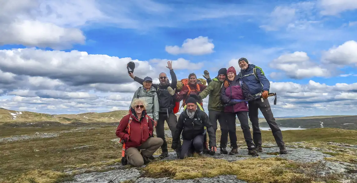 En grupp glada vandrare står tillsammans på ett fjäll med utsikt över ett vidsträckt landskap och blå himmel med moln.