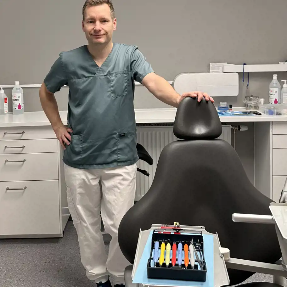 En tandläkare står leende bredvid en svart tandläkarstol i ett modernt behandlingsrum. På stolen framför syns ett uppdukat brickbord med färgglada tandläkarverktyg, och i bakgrunden finns vita skåp och rengöringsprodukter.