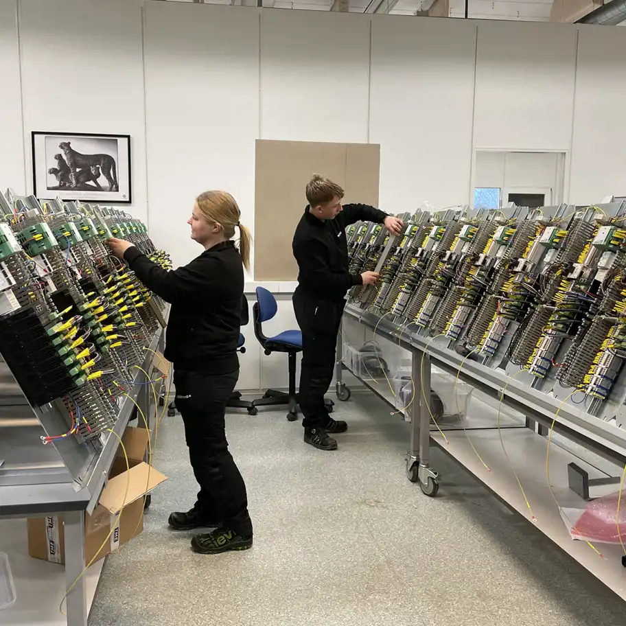 Två medarbetare arbetar fokuserat med att montera och koppla kablage i en rad elskåp på ett tekniskt produktionsgolv. Rummet är ljust och organiserat med moderna arbetsstationer.