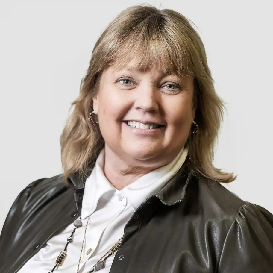 Närbild på en leende kvinna klädd i vit skjorta och mörk jacka, mot en ljus, neutral bakgrund.