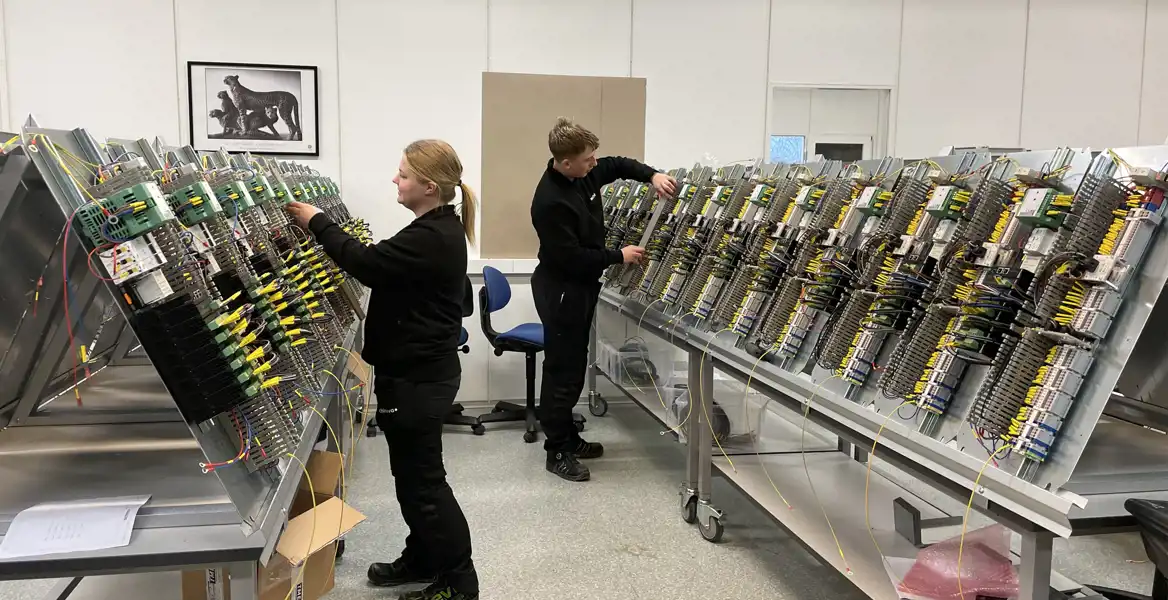Två medarbetare arbetar fokuserat med att montera och koppla kablage i en rad elskåp på ett tekniskt produktionsgolv. Rummet är ljust och organiserat med moderna arbetsstationer.