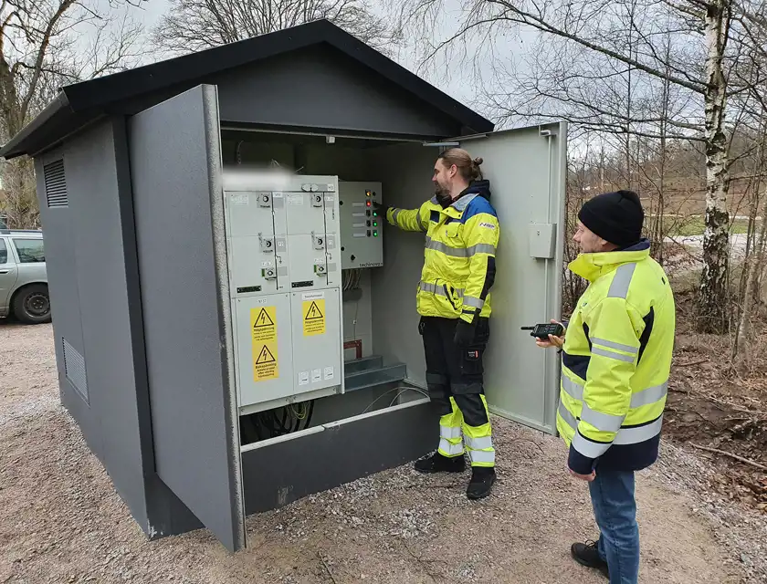 Två personer i reflexkläder arbetar vid ett öppet elskåp utomhus. Den ena trycker på knappar på kontrollpanelen medan den andra håller en kommunikationsradio. Omgivningen består av grus, träd och en grå himmel.