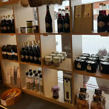 En välfylld trähylla i en gårdsbutik med lokalt producerade livsmedel som marmelad, saft, kryddor, kaffe och snacks. Produkterna är snyggt placerade i olika sektioner med handskrivna prislappar och charmiga förpackningar som skapar en hemtrevlig och genuin känsla.