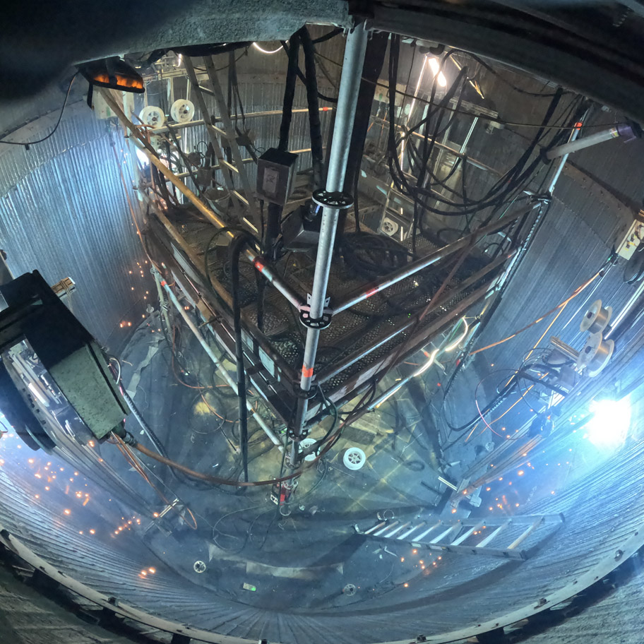 Inuti ett stort cylindriskt metallutrymme, fullt av avancerad teknisk utrustning och kablar. En plattform av metallstänger och nät är upphängd i mitten, omgiven av stegar, lampor och sladdar som hänger från väggarna. Ljuskällor och reflektioner skapar en futuristisk och intensiv arbetsmiljö.