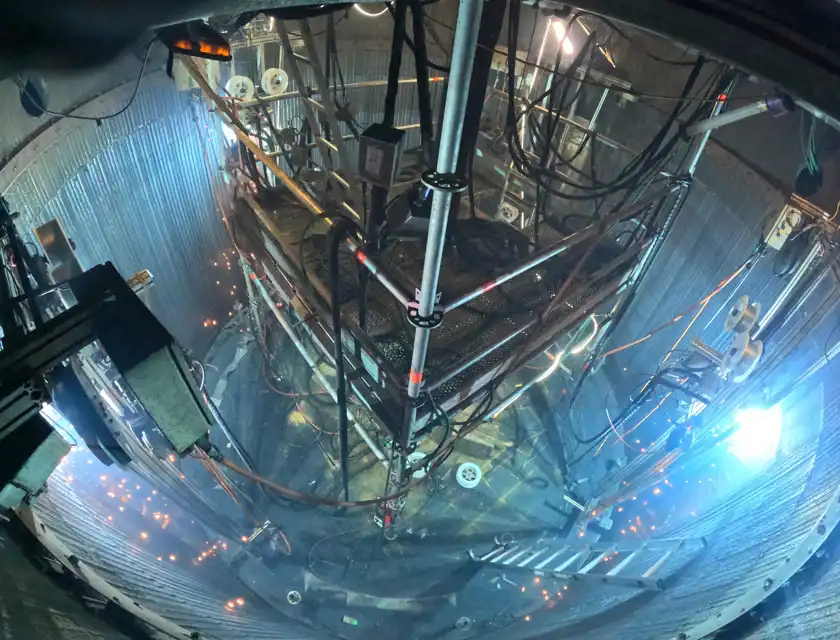 Inuti ett stort cylindriskt metallutrymme, fullt av avancerad teknisk utrustning och kablar. En plattform av metallstänger och nät är upphängd i mitten, omgiven av stegar, lampor och sladdar som hänger från väggarna. Ljuskällor och reflektioner skapar en futuristisk och intensiv arbetsmiljö.
