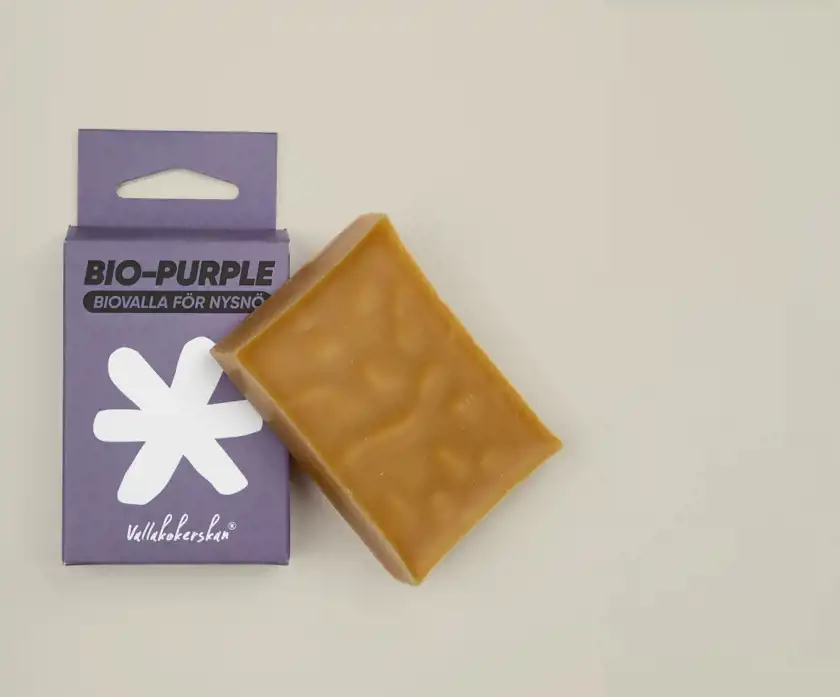 En bit biovalla i en gyllenbrun nyans ligger bredvid sin förpackning på en ljus bakgrund. Förpackningen är lila med en vit snöflingeliknande symbol och texten 'BIO-PURPLE – Biovalla för nysnö' samt varumärket 'Vallakokerskan' tryckt på framsidan.