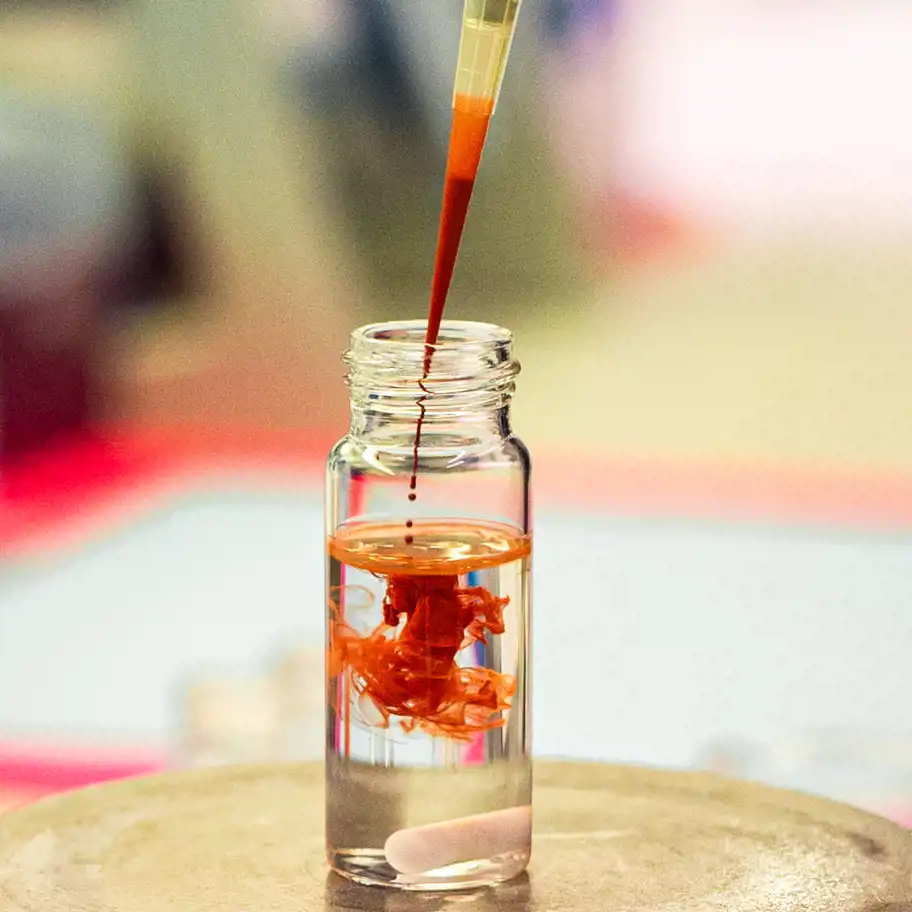 En närbild på ett genomskinligt glasrör som står på en laboratoriebänk. En pipett droppar en röd vätska ner i röret, vilket skapar virvlar i den klara vätskan i flaskan. I bakgrunden syns suddiga färger från laboratoriemiljön.