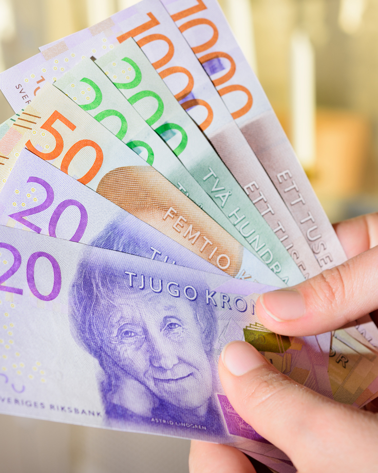 En hand som håller i en solfjäder av svenska sedlar i olika valörer, inklusive 20, 50, 100, 200, 500 och 1000 kronor. Sedlarna är färgglada och utspridda i en båge mot en oskarp bakgrund i inomhusmiljö.