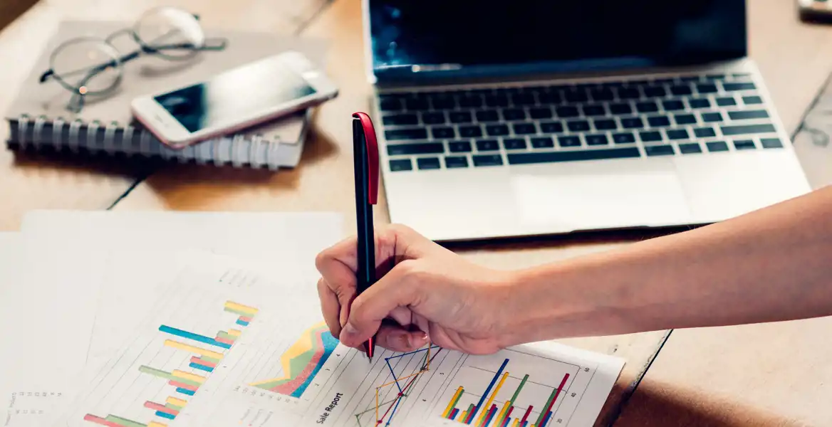 En person skriver med en penna på ett papper med färgglada diagram och grafer. På bordet ligger en bärbar dator, en smartphone, ett anteckningsblock och ett par glasögon. Arbetsmiljön är ljus och ser organiserad ut.