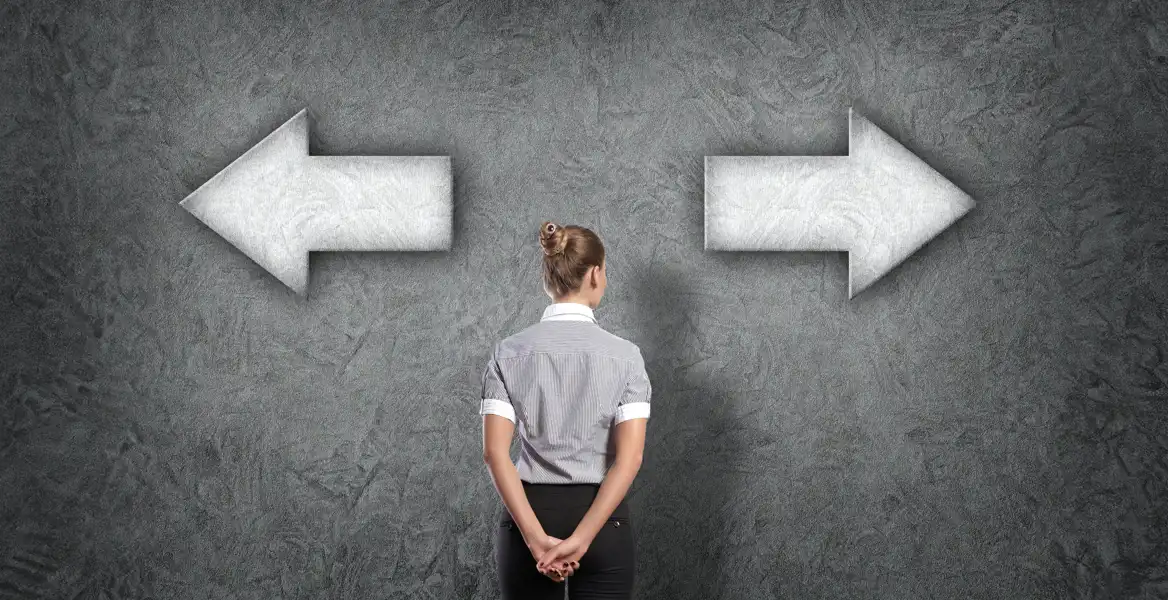 En kvinna med uppsatt hår står med ryggen mot kameran framför en mörk vägg. På väggen finns två stora pilar, en som pekar åt vänster och en åt höger. Hon står i mitten och ser ut att överväga vilken riktning hon ska ta. Symboliserar val eller beslutsfattande.