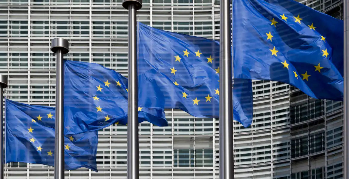 Fyra blå EU-flaggor med gula stjärnor vajar i vinden framför en modern byggnad med glasfasad och horisontella linjer, symboliserande Europeiska unionen och dess institutioner.