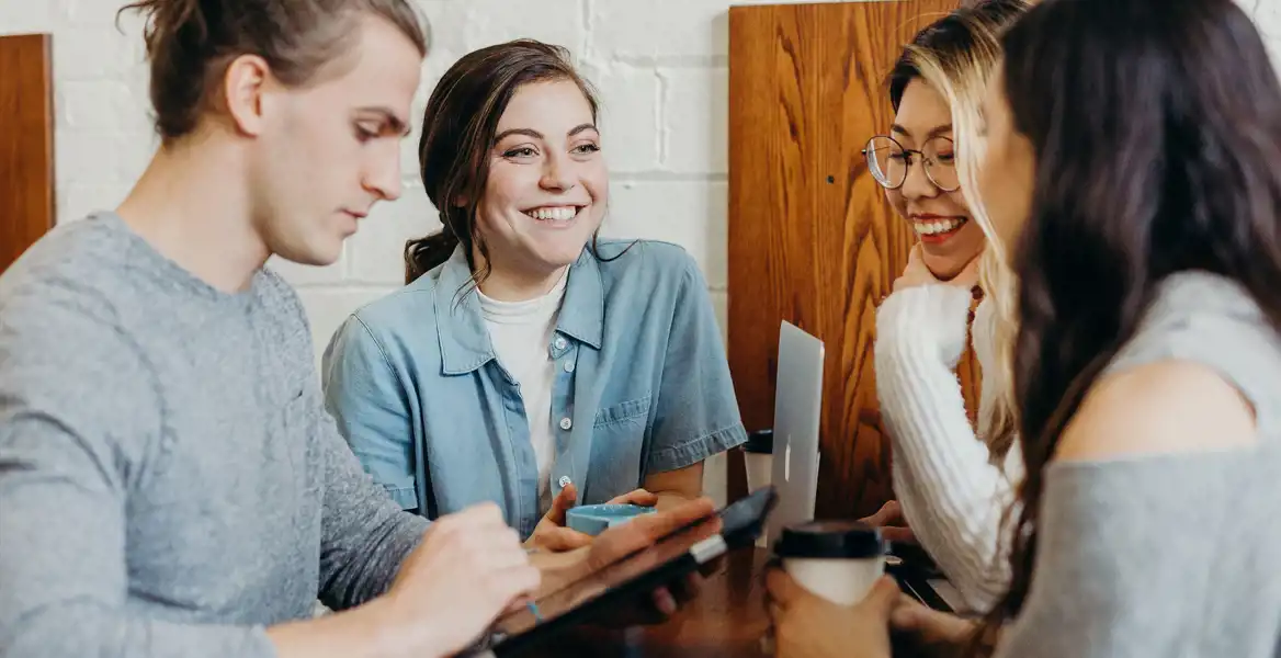 Fyra unga vuxna sitter runt ett bord och diskuterar med surfplatta, laptop och kaffe. De ser engagerade och glada ut. Bilden illustrerar en kreativ miljö där man kan utbyta idéer och träna på att presentera sin affärsidé – ett exempel på vad är en pitch i praktiken.