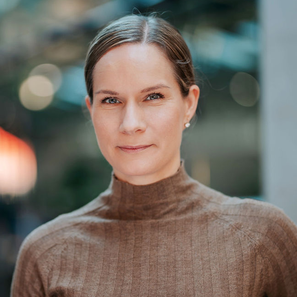 En kvinna står i en ljus inomhusmiljö. Hon bär en beige, ribbad polotröja och har håret uppsatt. Hon ser rakt in i kameran med ett lugnt uttryck och bakgrunden är mjukt suddig med varma toner.