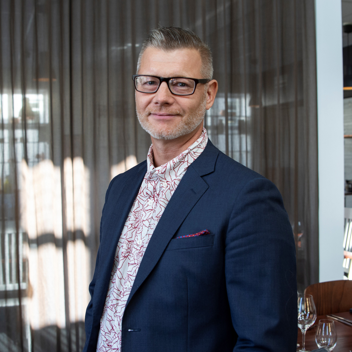 Man i kostym med glasögon står i en restaurangmiljö. Han ler mot kameran och har en mönstrad skjorta under sin mörkblå kavaj.